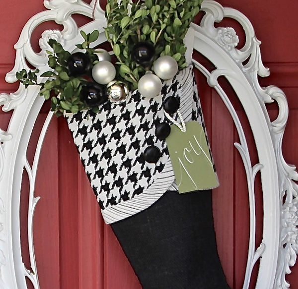 A closeup of a black and white Christmas stocking  with a  tag that reads "Joy", is shown  hanging int an ornate white frame on a front door