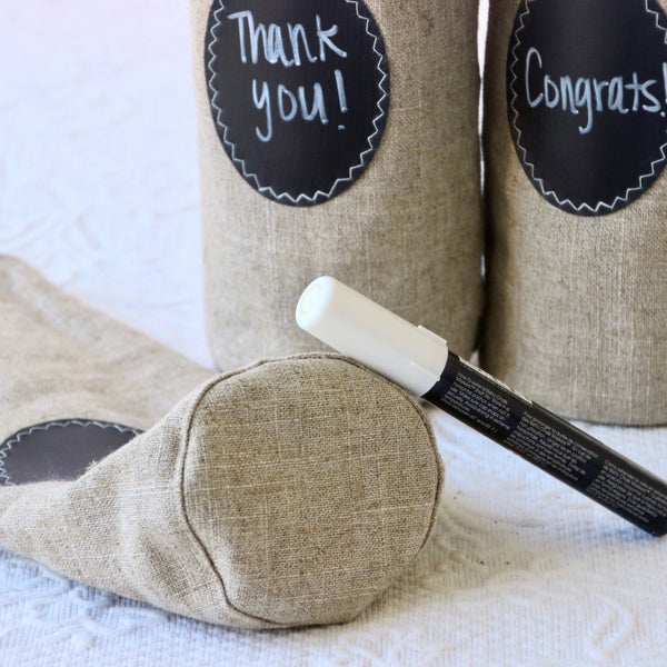 A Chalk marker is shown leaning against the base of a  linen wine bottle bag with two wine bottle bags behind it with messages on their chalk cloth labels